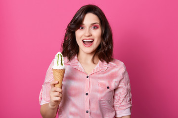 European funny young woman standing isolated over pink background in studio, opening her mouth widely with surprise, holding ice cream in one hand, being happy, wearing striped shirt. Food concept.