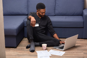 Image of serious pensive Afroamerican man staying at home, being confused with online services, holding credit card, looking at laptop, searching for information, having many documents around.