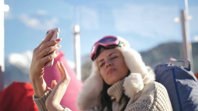 An attractive girl in a sweater and cap sitting in a outdoor cafe in the ski resort and taking a selfie on a smartphone.