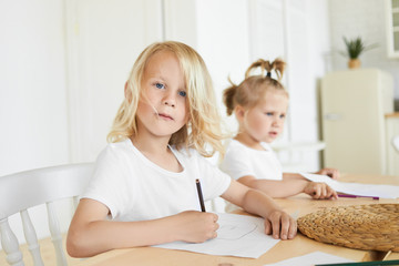 Fototapeta premium Two adorable Caucasian children doing homework together at wooden table. Cute seven year old boy with blond hair and blue eyes drawing at home with his little baby sister sitting in background