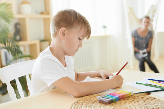 Busy boy of preteen age sitting at home with colorful plasticine on wooden table, using pencil, concentrated on creative process. Horizontal image of Caucasian little artist painting, doing homework