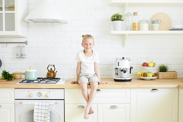 Indoor shot of cute handsome little boy with blonde hair bun sitting on counter in stylish kitchen, hanging down his bare feet, waiting for mother to cook breakfast in the morning before school