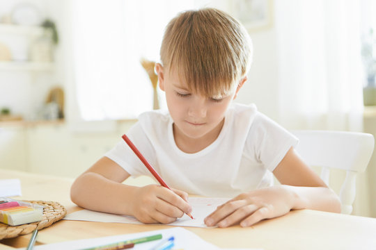 Serious focused schoolboy in white t-shirt entertaining himself indoors using red pencil drawing or sketching at wooden table isolated against stylish living room blurred interior background