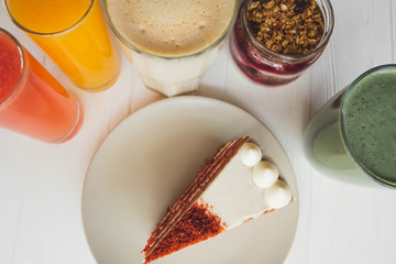 a red velvet cake on the background of glasses with various colored drinks (juice, coffee, smoothie, milkshake) standing on a white wooden table. A concept for a baker's dessert, a cafe, a restaurant