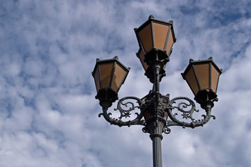 Vintage brown five-horn lantern in a classic performance with black plant and flower wrought iron swirls and corrugated relief support against the cloudy sky