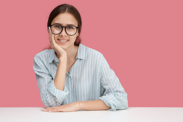 Beautiful happy young female employee wearing stylish blue shirt and spectacles enjoying working process, looking at camera with broad cheerful smile. People, lifestyle and occupation concept
