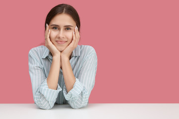 Charming friendly young Caucasian woman in stylish clothes sitting at desk with hands on her face,...