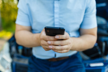 Smartphone in the hands of a man in a shirt shorts sitting on the front of the car with the hood open. Man are using their hands to ask for help because of a car accident or repair