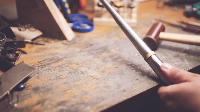 Close up of hands of jewelry maker bending a gold blank on road using a mallet hammer on workbench. Goldsmith making a ring in workshop.