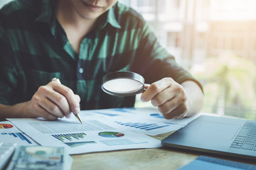 Business man using magnifying to review balance sheet annual with using laptop computer to...