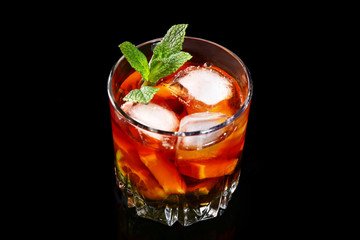 Glass of Dark Rum Cocktail with lime, orange, ice cubes and mint leaves on black mirror background.