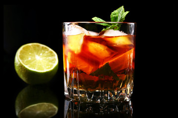 Glass of Dark Rum Cocktail with lime, orange, ice cubes and mint leaves on black mirror background.