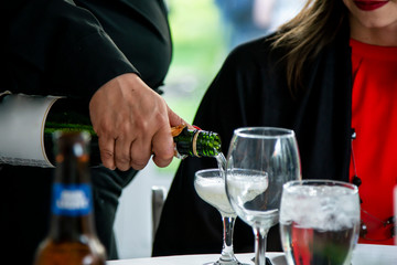 a male waiter is serving a woman with red lips and attire an alcoholic drink of champagne at a luxury wedding party.  the beverage bubbles up as  it is being poured in the fancy glass.  
