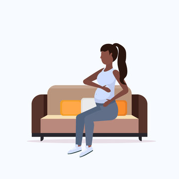 happy pregnant woman sitting on couch african american girl holding her bump girl pregnancy matherhood concept full length flat