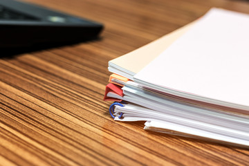 Pile of unfinished paperwork stacked in archive with plastic slide binder bars waiting to be managed and inspected for score and laptop nearby. Report papers stacks. Business and education concept.