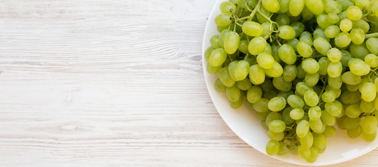 Green grape on a white plate on a white wooden surface, overhead view. Flat lay, top view, from above. Space for text.