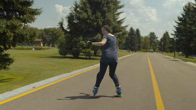 Stylish woman with beautiful long hair braided in many pigtails showing skill of backward sliding on roller blades along public park alley. Carefree female roller enjoying active leisure outdoors.