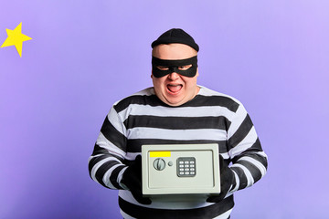 happy cheerful excited burglar in mask and striped clothes holding safety box isolated over blue....