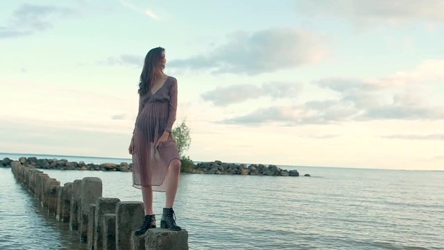 young attractive girl in transparent brown dress, black shoes standing on stone pillars sticking out of water at sunset. she straightens her hair and looks sexy at  camera. Evening picture of  model