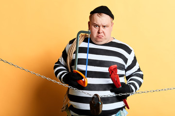 serious unhappy fat thief with different instrumnets standing over yellow background. close up...