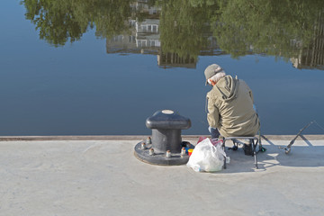 An old fisherman on the city quay with three fishing rods on the background of a blue river with houses and trees reflected in it. Concept: the life of elderly people in retirement.