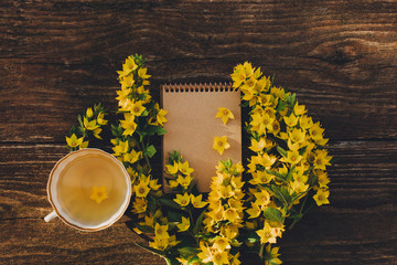 Blank notebook with yellow flowers and cup of tea on vintage wooden table. Flat lay, copy space.