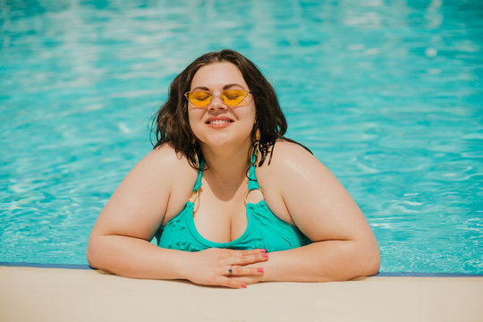Attractive busty curvy woman in yellow sunglasses and a blue swimsuit resting by the pool. Stylish accessories, fringe, fashion for plus size. Bodypositive, natural real beauty, resort, summer