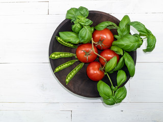 Tomatoes, basil and green pea pods in a dark wooden plate on a white wooden table