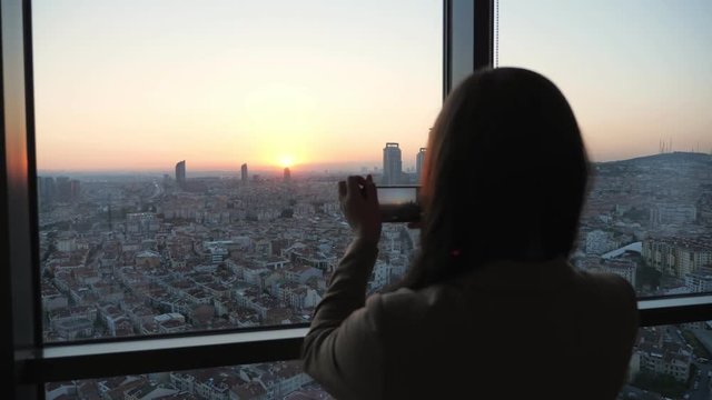 Silhouette of woman taking a pictures of panoramic city view at sunset on smartphone, focus on the city
