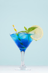 Blue curacao cocktail with ice cubes, lemon slice, yellow straw, green mint in luxury martini glass on white wood board and pastel mint color wall, vertical.