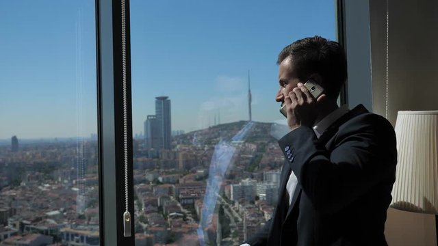 Young successful entrepreneur in office suit is talking mobile phone, side view. He is standing near the window with panoramic city view in his office at working day.