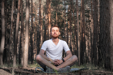 Young male person meditating in the forest. Modern man sits in the pine woods with closed eyes and...