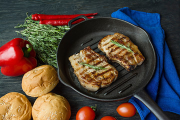 Grilled steak on a round grill pan, garnished with spices for meat, rosemary, greens and vegetables on a dark wooden background.