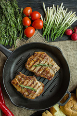 Grilled steak on a round grill pan, garnished with spices for meat, rosemary, greens and vegetables on a dark wooden background.