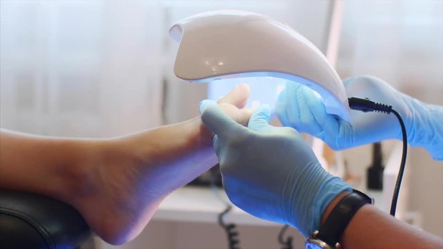 Professional pedicure is beauty salon. Pedicurist woman in gloves is holding UV lamp above the foot. She is drying shellac on client toes, foot closeup.