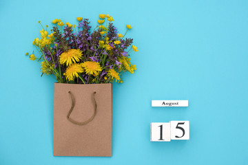 Wooden cubes calendar August 15 and field colorful rustic flowers in craft package on blue...