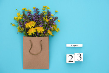 Wooden cubes calendar August 23 and field colorful rustic flowers in craft package on blue...
