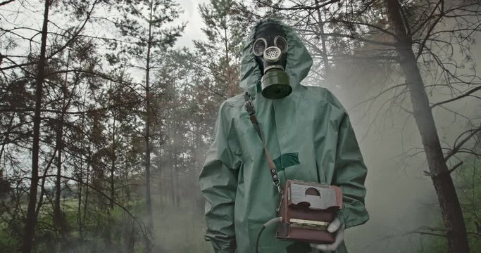 Low angle unrecognizable guy in hazmat suit and gas mask looking around while standing in cloud of smoke in middle of radioactive forest near Chernobyl.