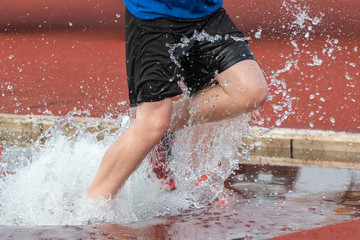 Runner in a steeple chase water bake on a running track.