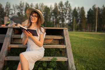 Naklejka premium Happy young woman sitting in the park. Smiling and holding a book in his hands. Sitting on a wooden bench. Sunset light