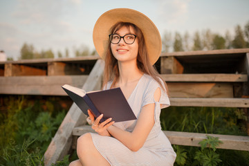 Obraz premium Happy young woman sitting in the park. Smiling and holding a book in his hands. Sitting on a wooden bench. Sunset light