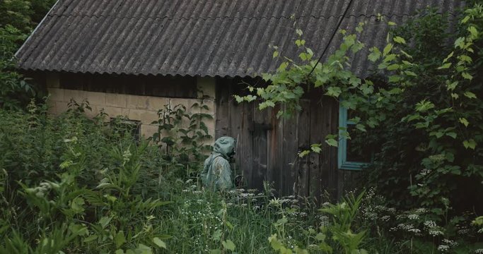 Anonymous male in hazmat suit and gas mask using Geiger counter on walls of desolated counter then turning around and searching for radiation on grass.
