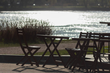 Fototapeta na wymiar Empty restaurant chairs and tables standing on the embankment of the river on a bright sunny summer day. Rest and eating in the open air, picnic. Summer vacation, relaxation and mood.