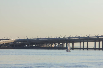 Fototapeta premium Blue wide river and the ring road above it. A lonely sailing yacht on the horizon is sailing on the sea against the background of the ring road passing over the water. Transportation concept.