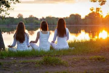 Yoga women dressed in white clothes doing yoga meditation at sunset by the lake,  sitting on a lush meadow. back view