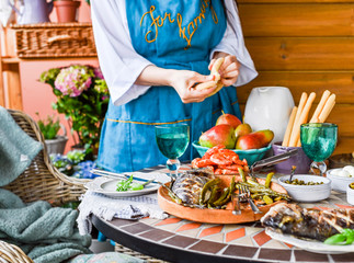 Fried fish in a dish in female hands. Dining table with different food and snacks. Summer lunch in the open air. Photo in bright colors. Woman in the frame at the dinner table.