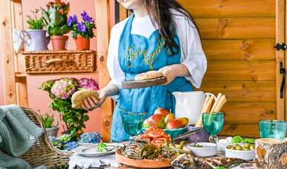 Fried fish in a dish in female hands. Dining table with different food and snacks. Summer lunch in the open air. Photo in bright colors. Woman in the frame at the dinner table.