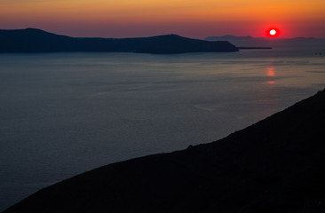 Fototapeta premium Oia santorini island greece. Natural coloured sunset. Dark red sky. Gold setting sun behind mountains. Calm sea reflecting sunlight. Beautiful colours. Thin clouds slightly visible.