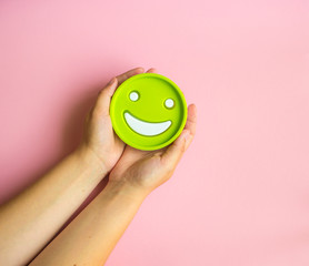 Smiley green face in woman's hands on pink background with copy space. Happy working day concept.