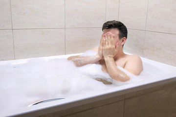 Adult man sitting in bath in the bathroom in foam, wipes face with his hands and relaxes in the evening at home.